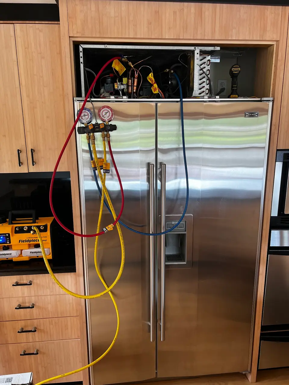 Refrigerant manifold gauges and sealed-system service equipment connected to a built-in side-by-side refrigerator with its top grille removed